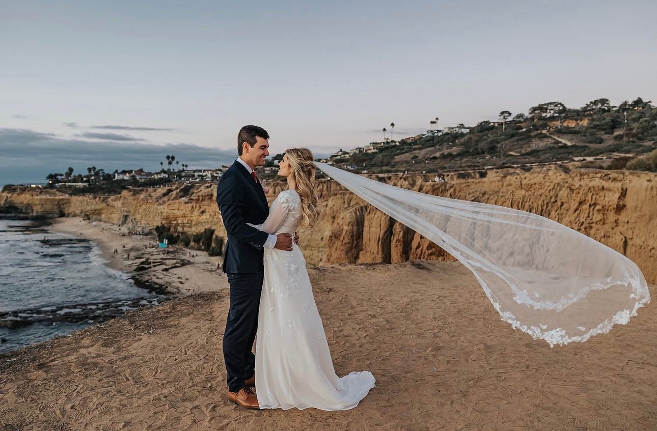 Bride and groom on a coastal cliff as her veil catches the wind